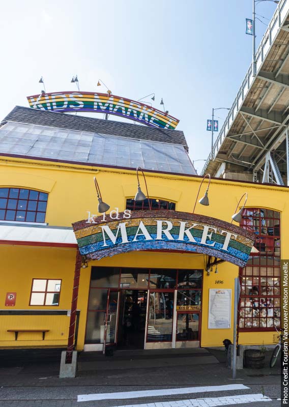 A yellow market building decorated with rainbows sits below a steel bridge.
