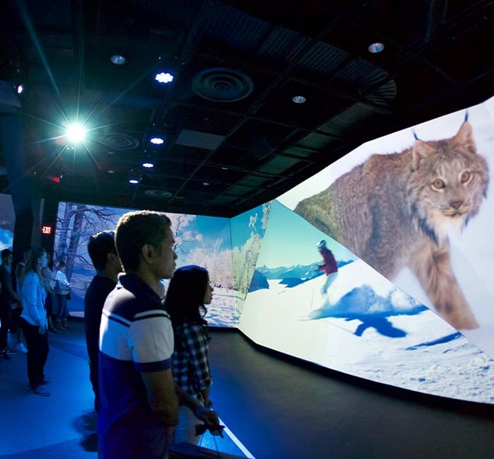 A group of people watch scenes of Canada on large screens.