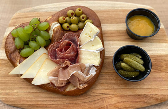 A charcuterie board served in a large pretzel with pickles and mustard.
