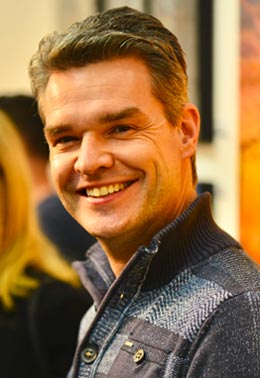 A smiling man with short brown hair, wearing a blue shirt