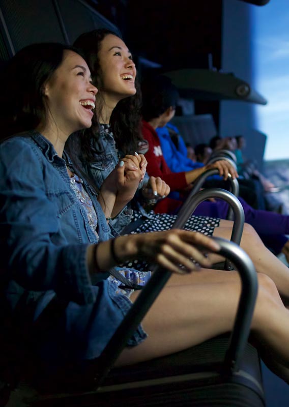Two smiling women hold on to their seats as the FlyOver Canada screen shows an outdoor scene
