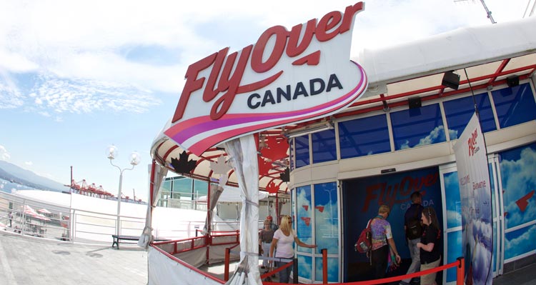 People enter the entrance of FlyOver Canada under sunny skies.