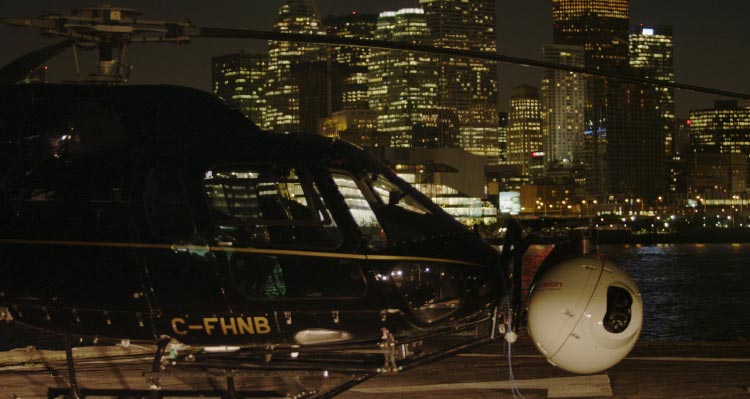 A helicopter with a video camera attached gets ready to take off with skyscrapers behind.