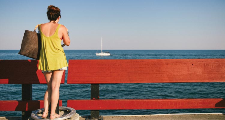 A person stands at a red fence looking out towards a sailboat on an expansive lake.
