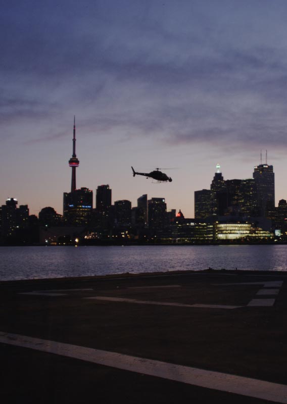 A helicopter flies over water between a helipad and the Toronto skyline at dusk.
