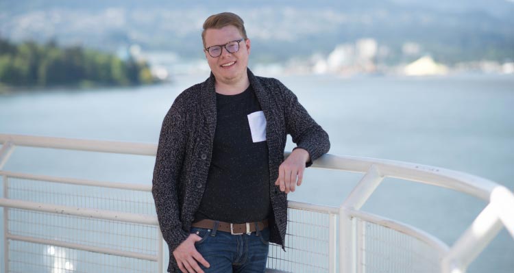 A man wearing glasses smiles as he leans on a railing by the sea.