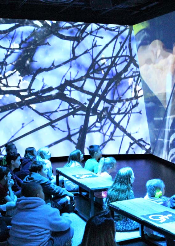 A group of children stand sit, watching a large projected video screen.