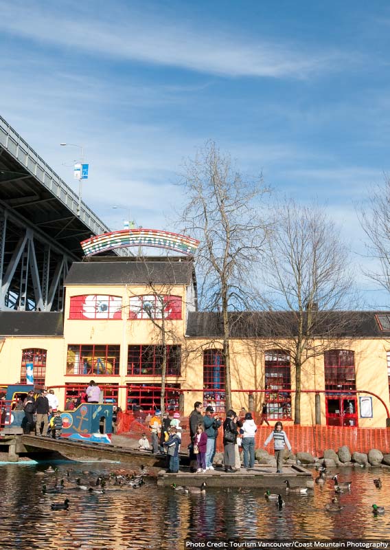 A yellow market building decorated with rainbows with families standing near ducks in the water