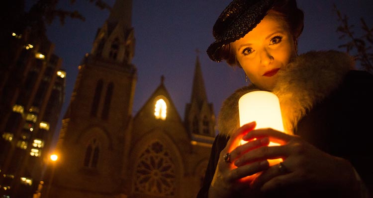 A woman holds a candle in front of a gothic cathedral at night time.