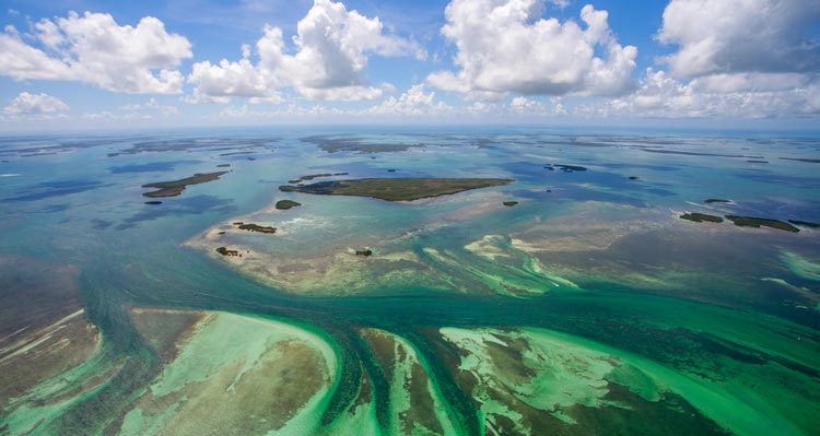 An aerial view of green tropical islands below a blue sky and fluffy white clouds.
