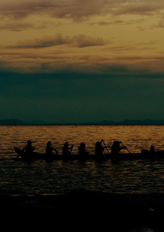 Six people paddle a large canoe near the seashore at sunset.