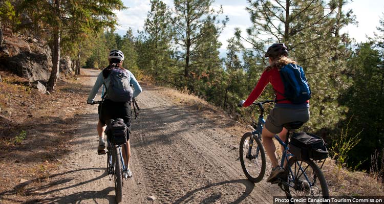 Two mountain bikers ride on a dirt pathway along a forested hillside.
