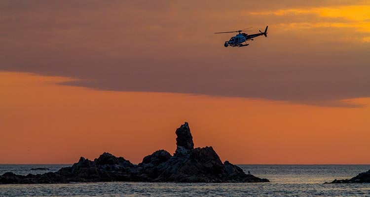 A helicopter flies over a rocky outcrop from the ocean.