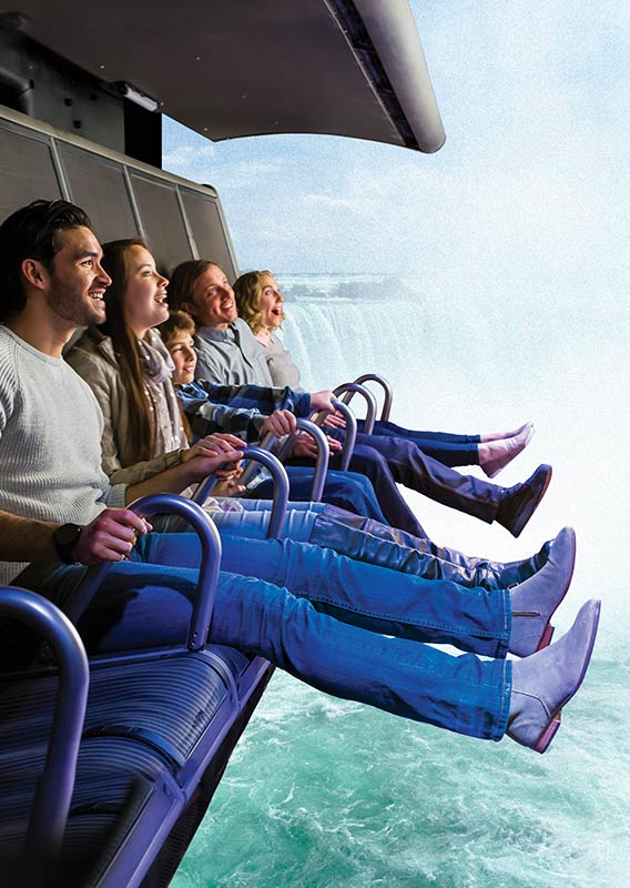 A group of people sit on the FlyOver Canada Flight Ride, with a view of Niagara Falls behind them.