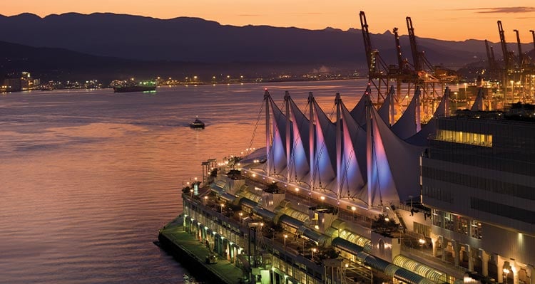 An aerial view of a white building, lit up at dusk, next to a wide harbour.