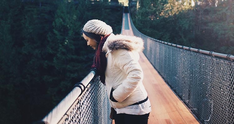 A woman stands on a narrow suspension bridge, looking down into a canyon.