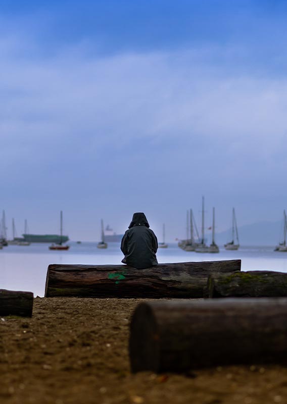 A person sits on a log bench on a beach looking out at a harbour.