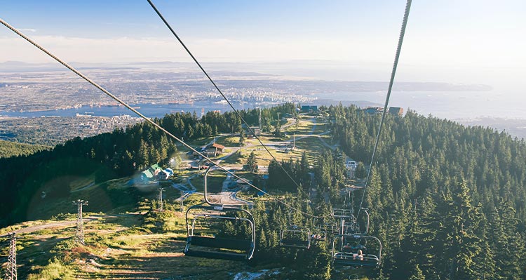 People ride the chair lift up the mountain in the summer, city in the distance.