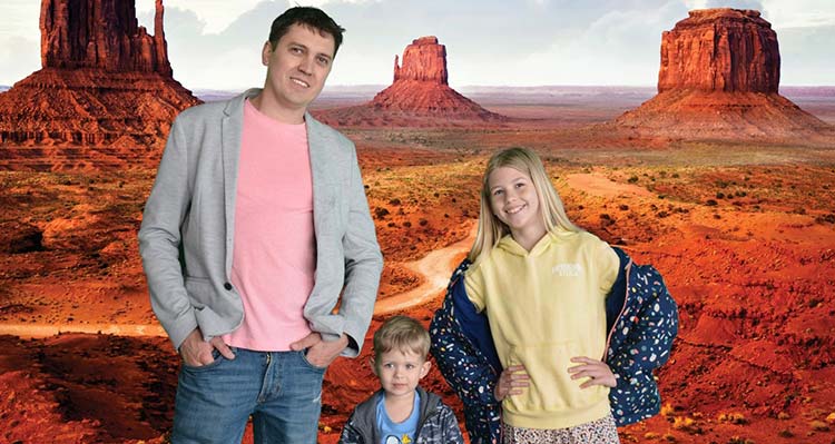 A man, a young girl and a little boy pose in front of the Real Wild West FlyOver Canada background.