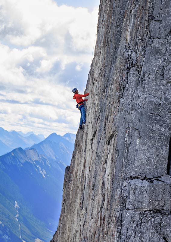 A man in a red jacket rocks climbs alone up a steep rockface.