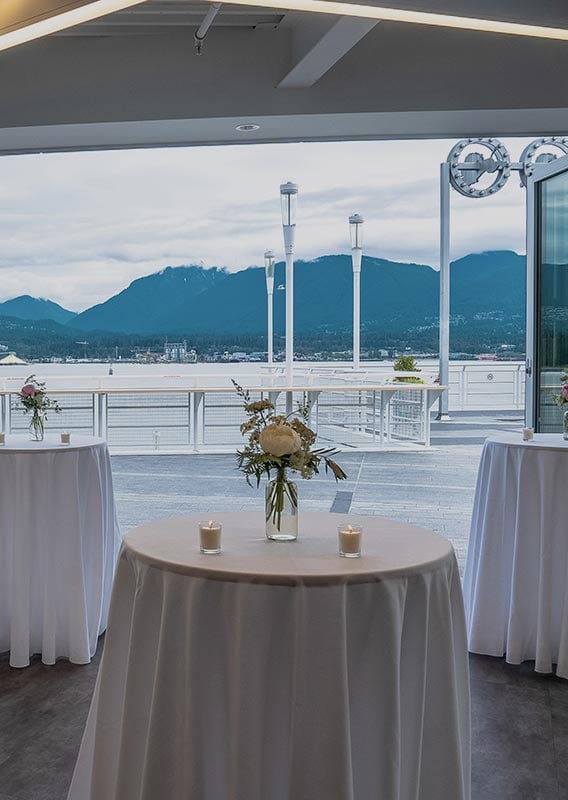 A set of small tables set up for an event in a building with wide open doors overlooking the water.