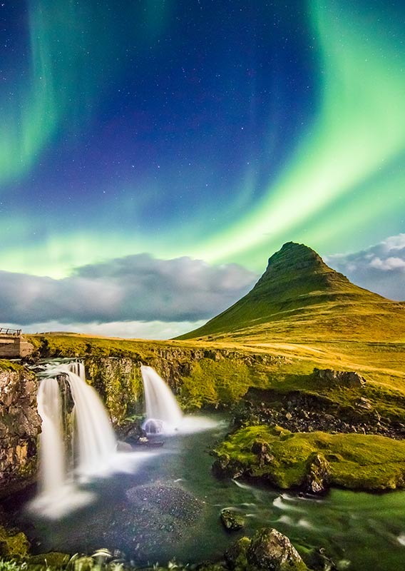 A waterfall rushing below a tall mountain and northern lights.